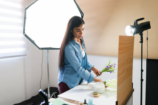 Young female photographer arranging prop flowers and a cosmetic product for a creative flat lay still life setup