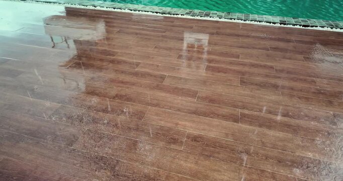 Slow motion: heavy rain falls on a wet, wood patterned tiled floor next to a swimming pool, creating ripples and splashes while reflecting the image of a nearby building.