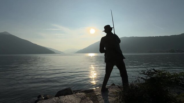 Switzerland. Fisherman in silhouette stands on rocky shore casting fishing rod into calm lake at sunrise, with mountains and trees framing the serene landscape