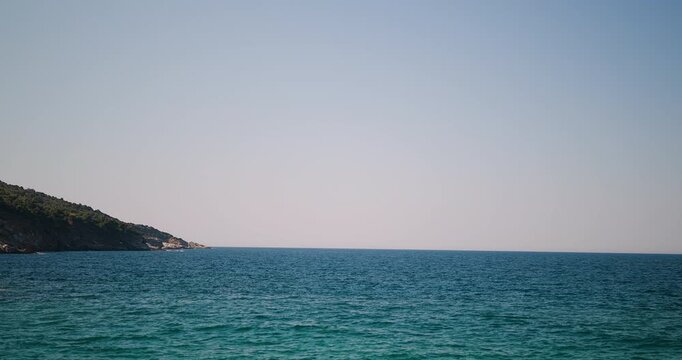 Serene Coastal Landscape with Clear Turquoise Waters Gently Washing Against a Tree-Covered Rocky Shoreline Under a Bright Blue Sky, Capturing the Tranquil Beauty of Nature and the Vast Ocean Horizon