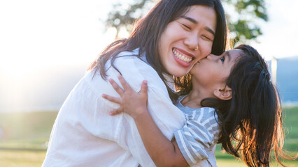 Fototapeta premium Smiling Asian mother holding her young daughter who is giving her a loving kiss on the cheek. Concept of happy family lifestyle, self-care and wellness.