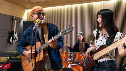 Fototapeta premium Happy Asian music band performing together with acoustic guitar, bass, and drums in a recording studio. Concept of joyful rehearsal, teamwork and music passion.