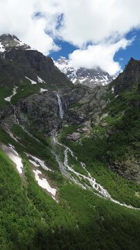 Vertical drone footage approaching Shdugra waterfall in Mount Ushba's rocky gorge, Georgia. Snow-capped summit, melting glacier and river below, under blue sky. Original unedited 4K footage.