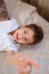 Fototapeta premium A young child in a white shirt and suspenders lies on a floral patterned bed cover, smiling and pointing playfully with one hand toward the camera