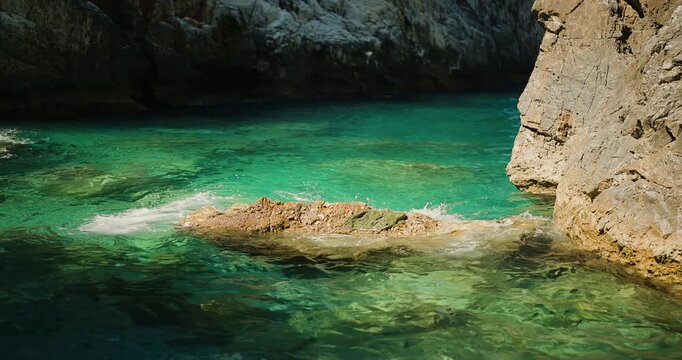 Gentle turquoise waves crash against sunlit rocks and rugged cliffs, creating a serene and beautiful natural seascape, perfect for relaxation and nature documentaries.