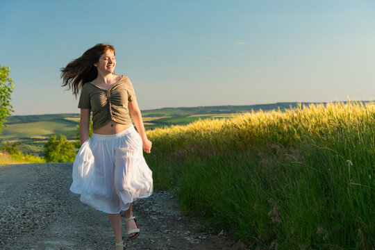 Happy young woman walking in white skirt on countryside road during golden hour