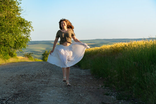 Cheerful young woman in white skirt walking on country road at sunset