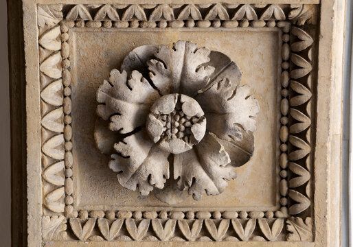 Antique carved stone flower rosette in a square ornamental frame, front-view close-up of weathered architectural relief with symmetrical leaf border, decorative historic masonry detail