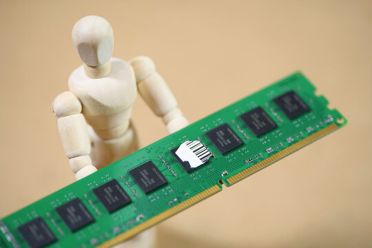 Wooden figurine holding a green desktop RAM module on a brown background, symbolic close-up of computer memory upgrade, chip shortage, AI hardware demand and rising component prices