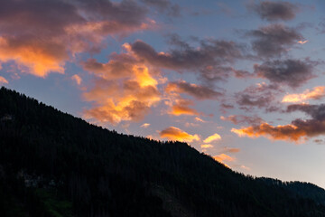 Obraz premium Dramatic alpine sunset cloudscape over silhouetted mountain ridges and pine forest, featuring glowing orange clouds against a deep blue sky, Dolomites, South Tyrol, Italy