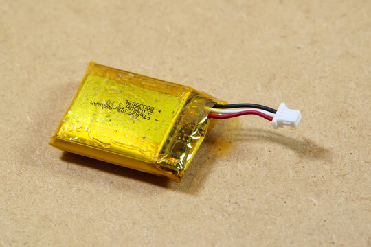 Swollen yellow lithium-ion pouch battery with three-wire connector on a brown surface, damaged rechargeable cell posing fire, overheating and e-waste disposal hazards, close-up with copy space