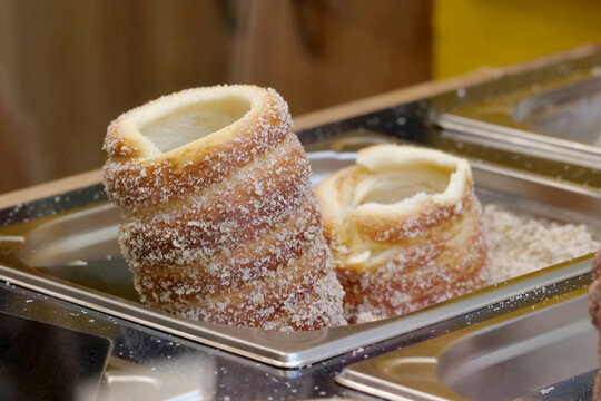 Fresh sugar-coated chimney cakes standing in a stainless steel tray at a bakery or street food stall, close-up of hollow spiral pastries with crisp golden crust, crumbs and no people
