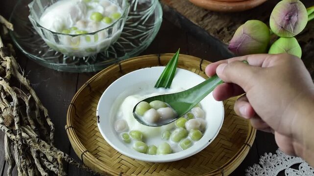 Top view of a hand holding a spoon scooping traditional Thai dessert Bua Loi, colorful rice flour balls in warm sweet coconut milk, served in a white bowl on a rustic wicker tray.