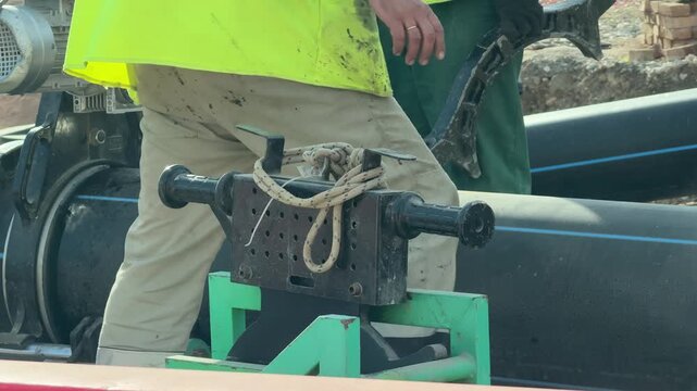 Construction workers welding large black plastic pipes on site. Close up shot captures manual labor and professional pipe fusing equipment in operation.