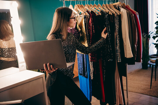 Fashion stylist reviewing wardrobe choices on laptop while selecting sequin dresses from a clothing rack in a dressing room