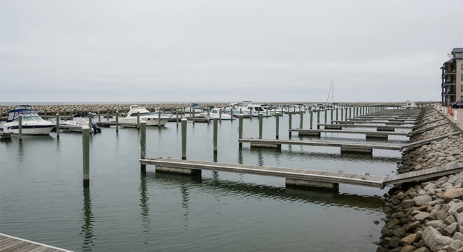 Empty wooden boat slips and floating docks at a coastal marina under overcast sky with motorboats moored near a rocky breakwater and nautical harbor infrastructure.