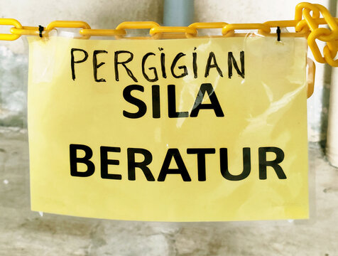 An editorial healthcare photograph of a yellow sign with black text in Malay reading PERGIGIAN SILA BERATUR, meaning Dental, please queue. The sign is attached to a yellow plastic chain in a clinic se