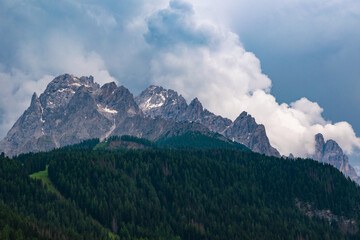 Obraz premium Majestic panoramic view of rugged mountain peaks rising above dense pine forests under a dramatic, cloudy sky in the Dolomites