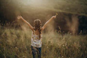 Naklejka premium Little girl standing with raised arms in a field at sunset, rear view