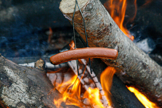 Hot dog roasting over a crackling campfire with glowing embers and smoke