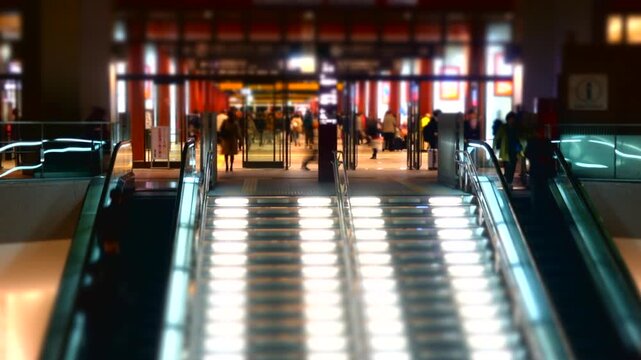 Tilt-shift timelapse footage of a busy railway station entrance in Japan at night, featuring illuminated escalators, commuters walking through the station, and vibrant urban lighting. The miniature ef