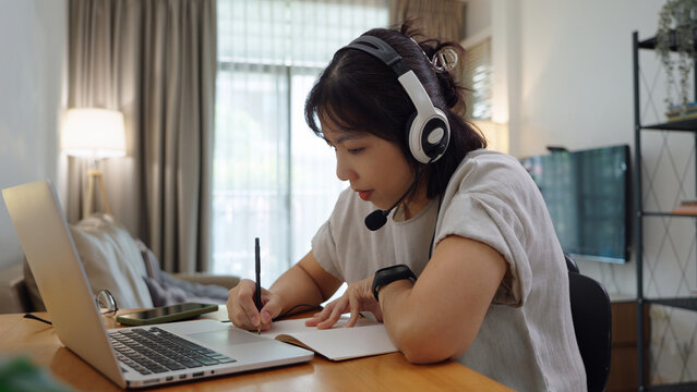 Asian college female student writing notes and doing homework while attending an online class with a laptop and headset at home.