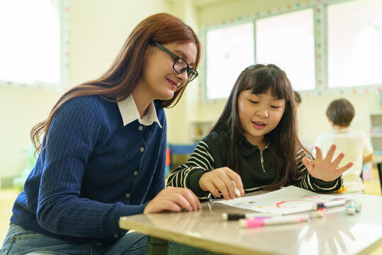 Caucasian female teacher helping Asian girl during an art lesson with drawing and coloring activities in the classroom.