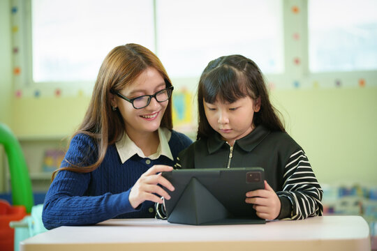Caucasian female teacher and Asian girl using a digital tablet together during a technology lesson in the classroom.