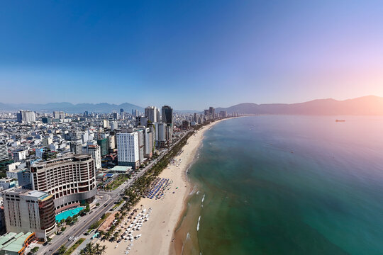 Expansive aerial view of Da Nang: Long sandy beach, modern high-rises, clear turquoise ocean, distant mountains, sunny sky