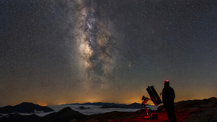 Fototapeta premium Amateur astronomer with telescope and red headlamp watching the Milky Way above a sea of clouds