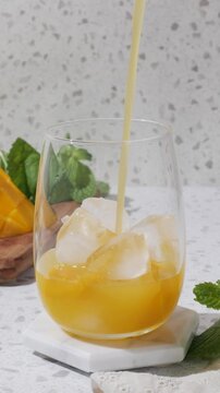 Summer mango drink mocktail with ice and mint on a white coaster, styled in bright sunlight on a terrazzo countertop with fresh mango and clean copy space. A clear rounded glass holds a golden mango