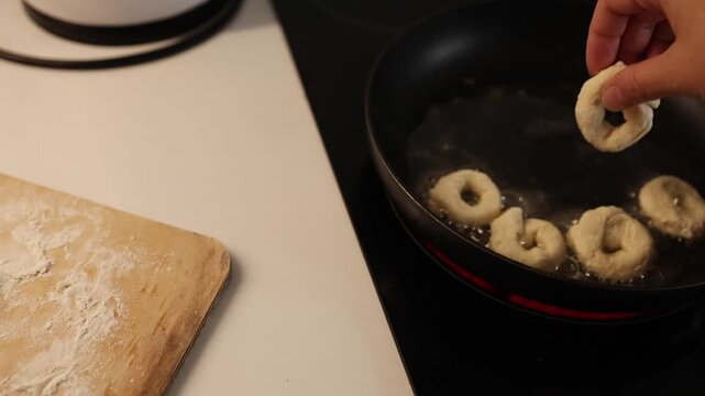 A person is frying donuts in a pan with hot oil, flipping them to achieve a golden‑brown crust, in a kitchen setting, home cooking activity, comfort food preparation.