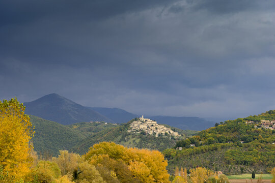 Scenic autumn landscape of Lago di Piediluco area  featuring a historic hilltop village surrounded by mountains and colorful yellow trees under a dramatic stormy sky