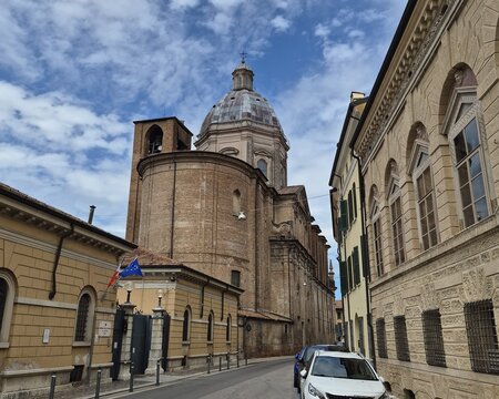 View of San Barnaba church from Via Poma in Mantua, showing its brick apse, majestic dome, and historic buildings under a cloudy blue sky.