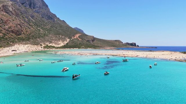 Aerial drone view of Balos Lagoon with crowds on the white-sand beach below rugged mountains, Balos, Greece