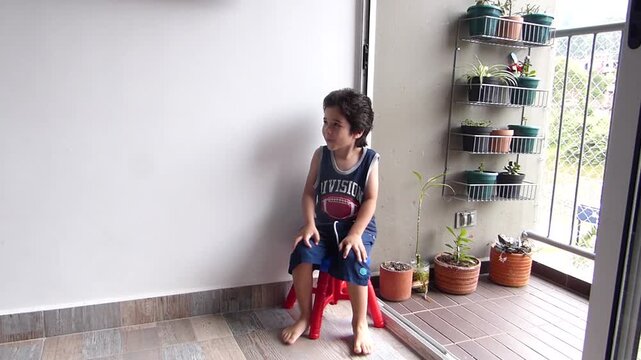 Happy little hispanic boy smiling on a red plastic stool while sitting on an apartment balcony with potted plants, portrait look at camera
