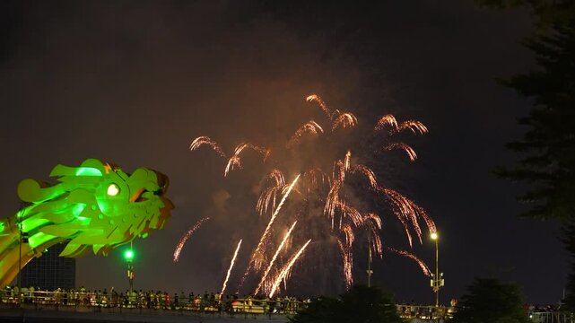 Bright explosions light up the night sky above Da Nang Dragon Bridge and Han River in Vietnam. Crowds gather to enjoy the amazing fireworks show