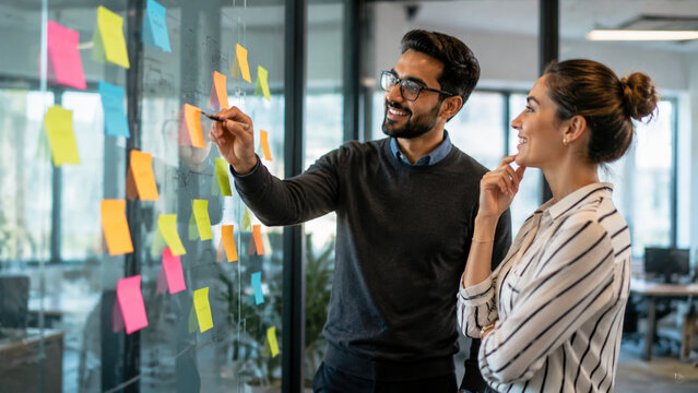 Professional man and woman collaborating on a project with colorful sticky notes on a glass wall in a modern office