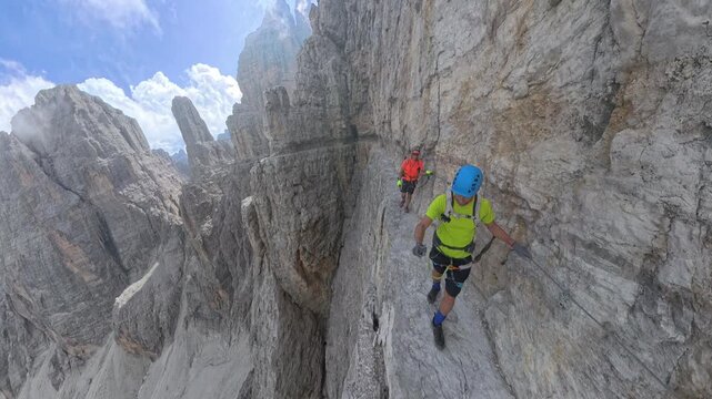 Two hikers crossing an exposed ledge on Via Ferrata Bocchette Centrali with dramatic drops below, Brenta Dolomites
