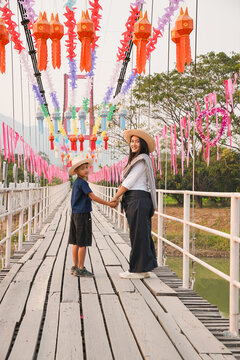 A happy Asian mother and son holding hands on a wooden bridge decorated with colorful lanterns during Songkran festival or Thai New Year travel, local village lifestyle outdoor.