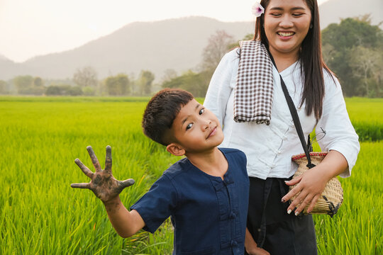 A playful Asian boy tilting his head and showing his muddy hand to the camera with a smiling mother in a green rice field background, authentic rural family lifestyle outdoor.