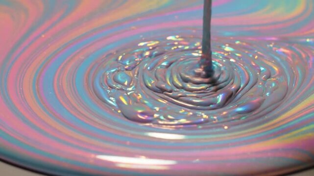 Macro close-up of thick iridescent liquid pouring and swirling into a container showing vibrant pastel pink, blue, and yellow colors with a metallic pearlescent finish.