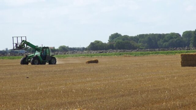 Tractor harvesting an agricultural field by baling straw into large round hay bales after crop harvest.