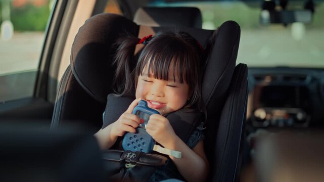 Happy Asian toddler girl smiling while sitting in safety car seat, cute little child holding a toy during family road trip vacation for child safety, parenting travel, automobile insurance, family