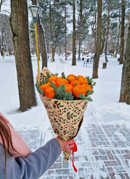 Young woman in a grey coat holds a unique bouquet of vibrant tangerines and pine sprigs as walks in the snowy park, creating a cozy winter scene. Festive holiday warmth and seasonal gift concept