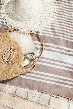 Summer flat lay of round straw bag, bracelet of seashell, clear glass water bottle, white sun hat on beach towel on wood. Vacation, travel, resort, and summer fashion concepts. Neutral beige color.