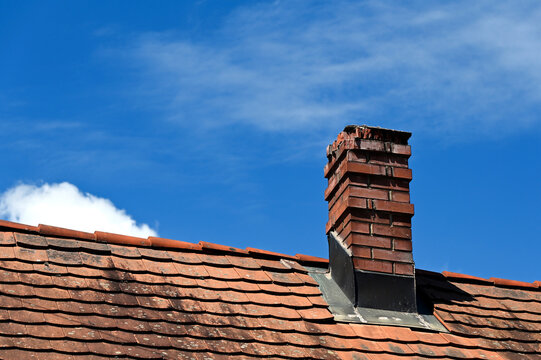 damaged clay brick chimney with weathered and spalling surface. clay tile roofing. gray metal flashing. strong shadow. blue sky background. summer scene. building, home maintenance and upkeep concept