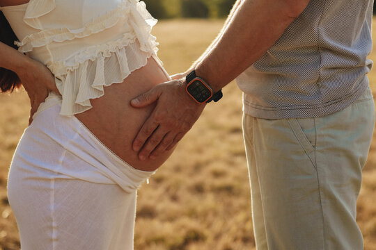 Close up of a husband placing his hand on his pregnant wife's bare belly outdoors at sunset, maternity and family support concept