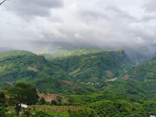 Misty mountain landscape with rainbow over green hills and cloudy sky. © Sheikh