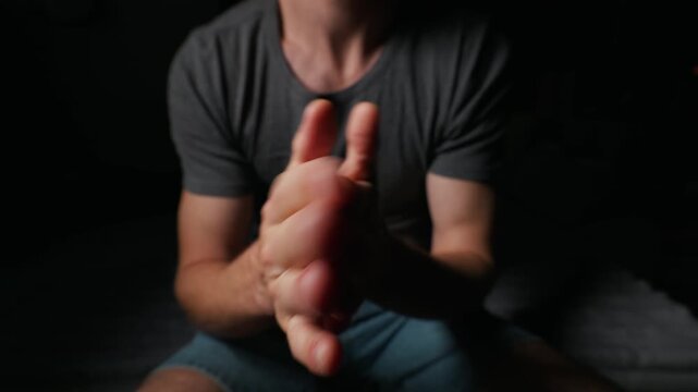 A young man sits in a shadowy room while anxiously rubbing his hands together in low light. Concept of anxiety, mental health, anticipation, nervous tension, stress, and expectation.